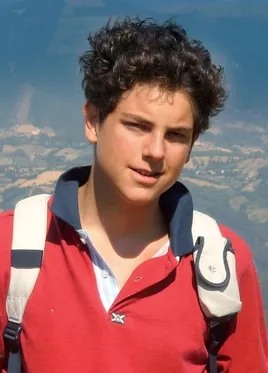 Carlo Acutis vistiendo una camisa polo roja de cuello azul marino, trae una mochila a los hombros, de fondo se ve un paisaje toscano