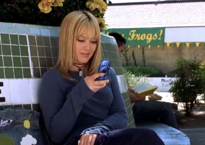 Frame de la la película La Nueva Cenicienta. La actriz  Hillary Duff envía un mensaje de texto con un flip phone mientras está sentada en una fuente.
