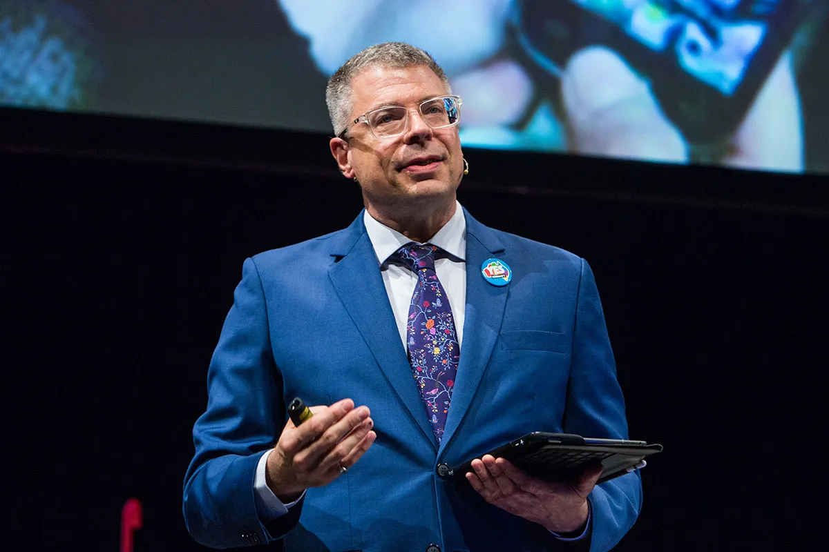 Fotoretrato de Mark Pesce, viste un traje sastre azul marino con una corbata con motivos florales, usa lentes cuadrados de marco transparente y sostiene una tableta con teclado, está dando una conferencia.